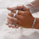 Two hands covered in sand clasp together. The person wears multiple silver rings and bracelets, with fine sand falling between their fingers onto a sandy surface.