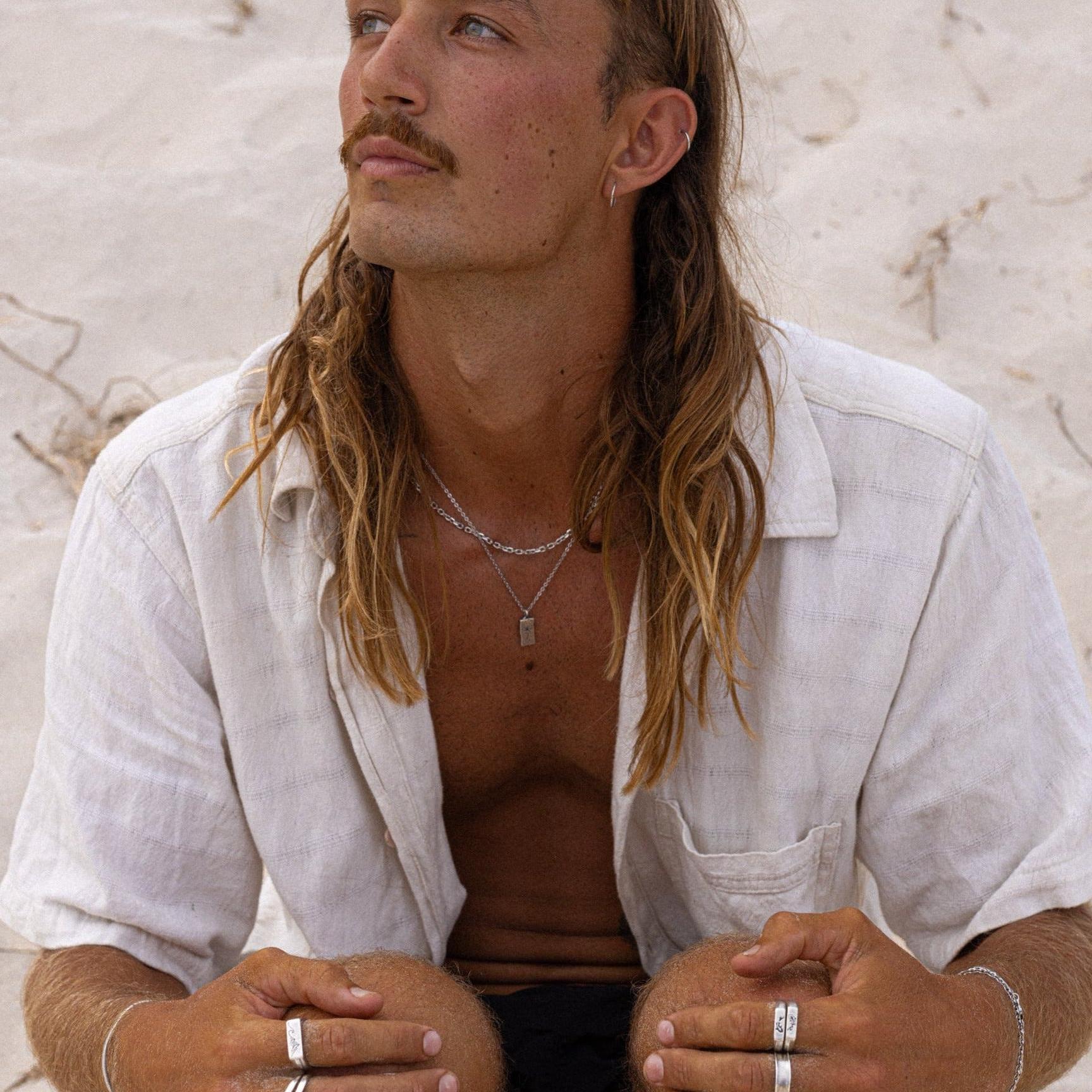 A man with long hair and a mustache sits on sandy ground, wearing a light-colored, short-sleeved shirt, multiple rings, and layered necklaces, looking slightly upward with a thoughtful expression. v2