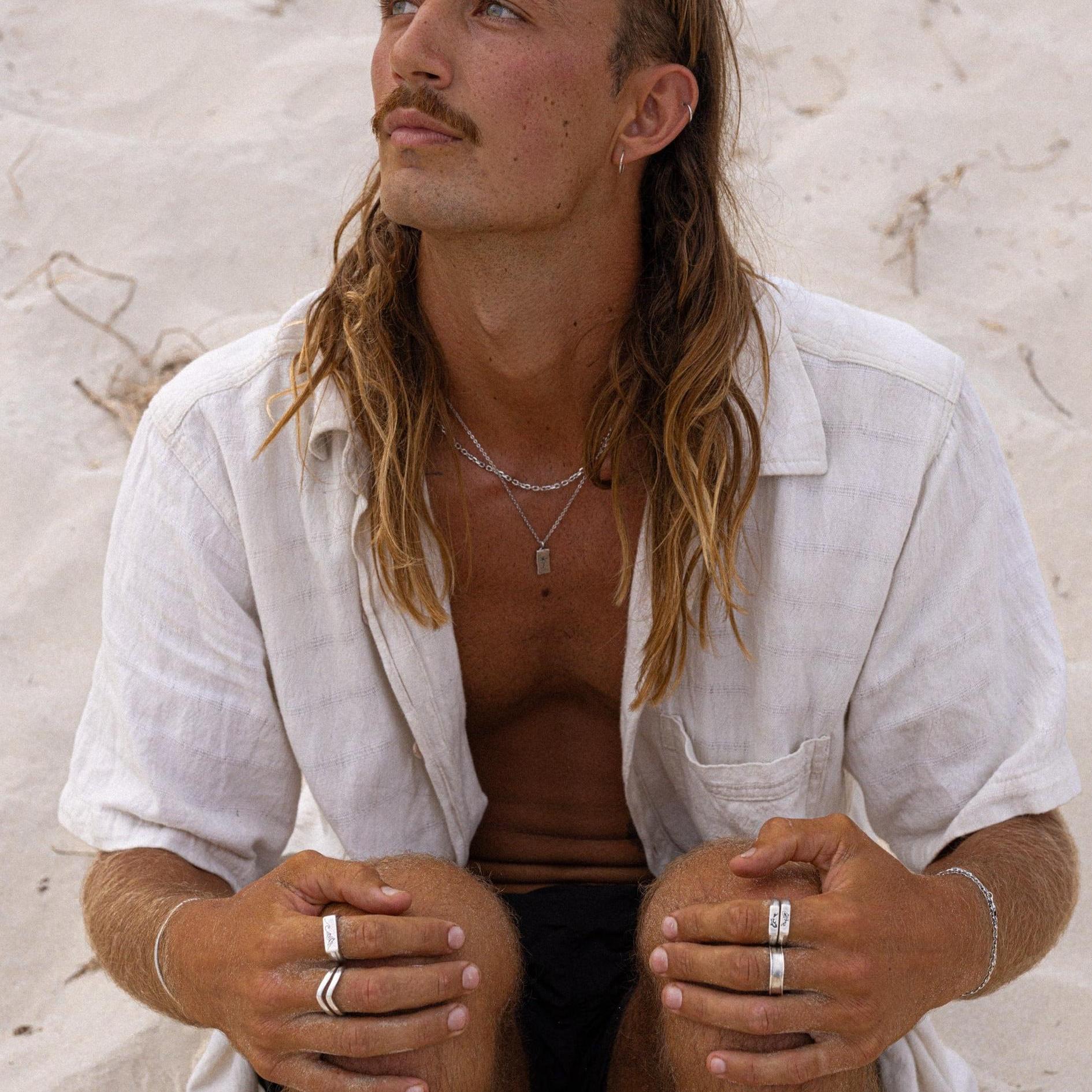A man with long hair and a mustache sits on sandy ground, wearing a light-colored, short-sleeved shirt, multiple rings, and layered necklaces, looking slightly upward with a thoughtful expression. v4