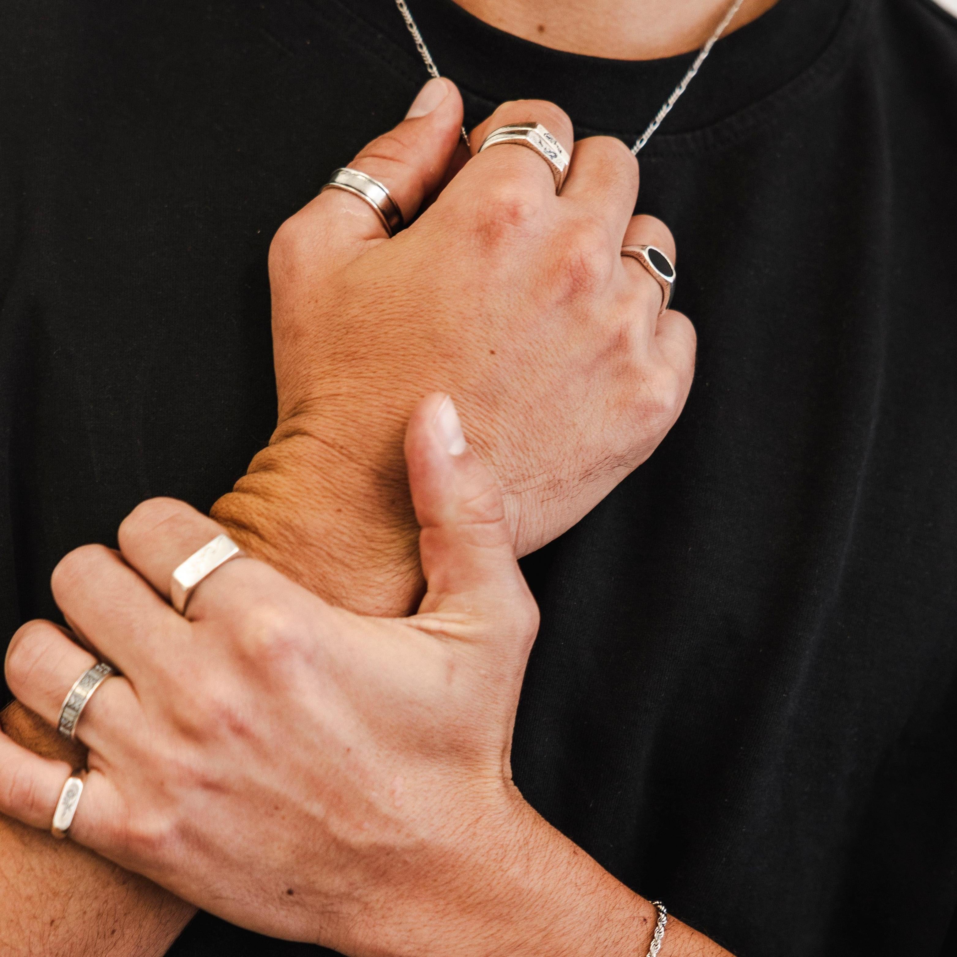 A person in a black t-shirt holds their wrist, showing multiple silver rings on their fingers, a chain bracelet, and a necklace. Only the hands, part of the neck, and torso are visible.