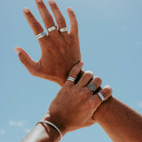 Two hands adorned with multiple silver rings and a bracelet are raised against a bright blue sky. The fingers are slightly spread, showcasing the jewelry in natural sunlight.