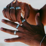Two hands with medium brown skin tone wearing multiple silver rings and a silver bracelet, posed against a blurred light background, possibly outdoors in natural light.