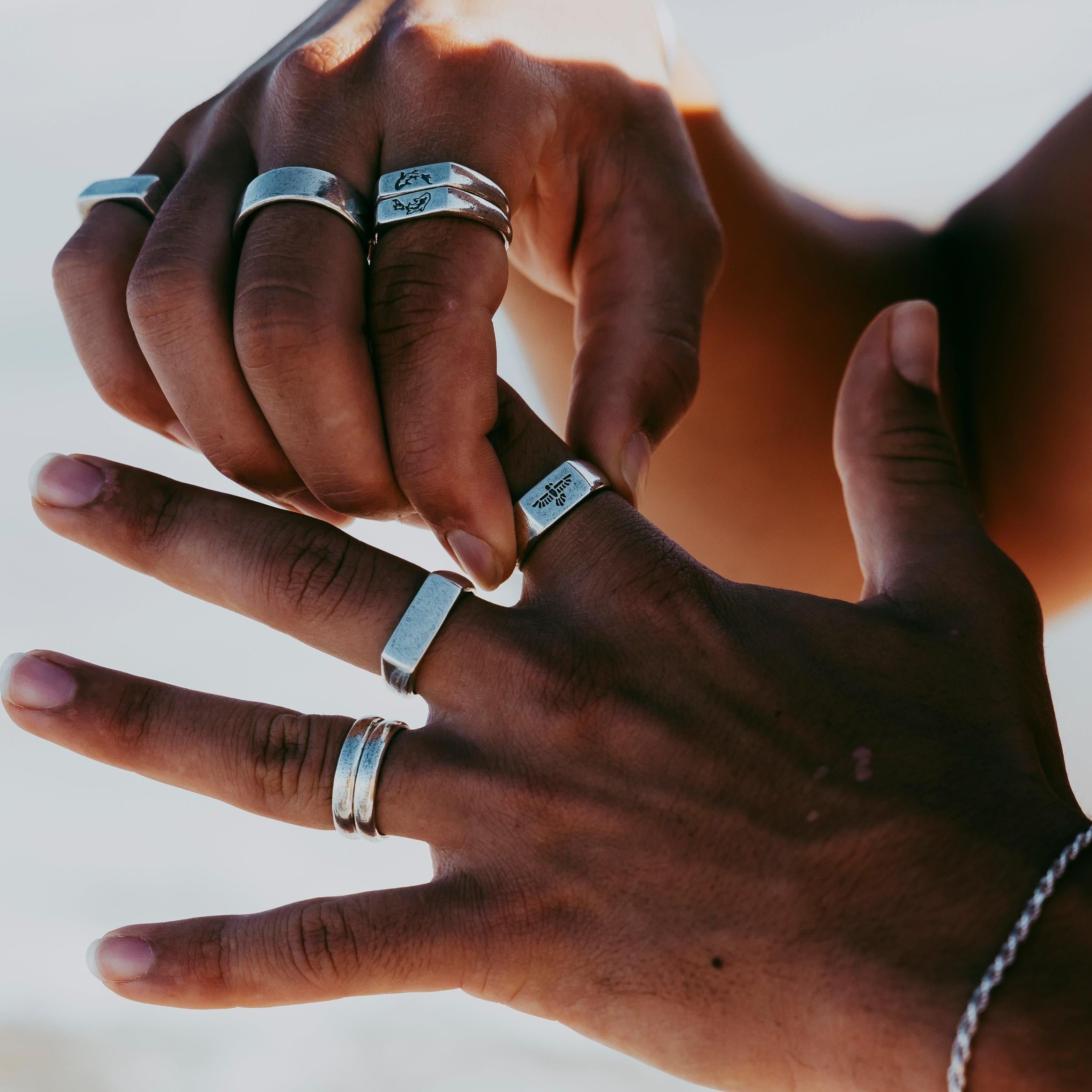 Two hands with medium brown skin tone wearing multiple silver rings and a silver bracelet, posed against a blurred light background, possibly outdoors in natural light.