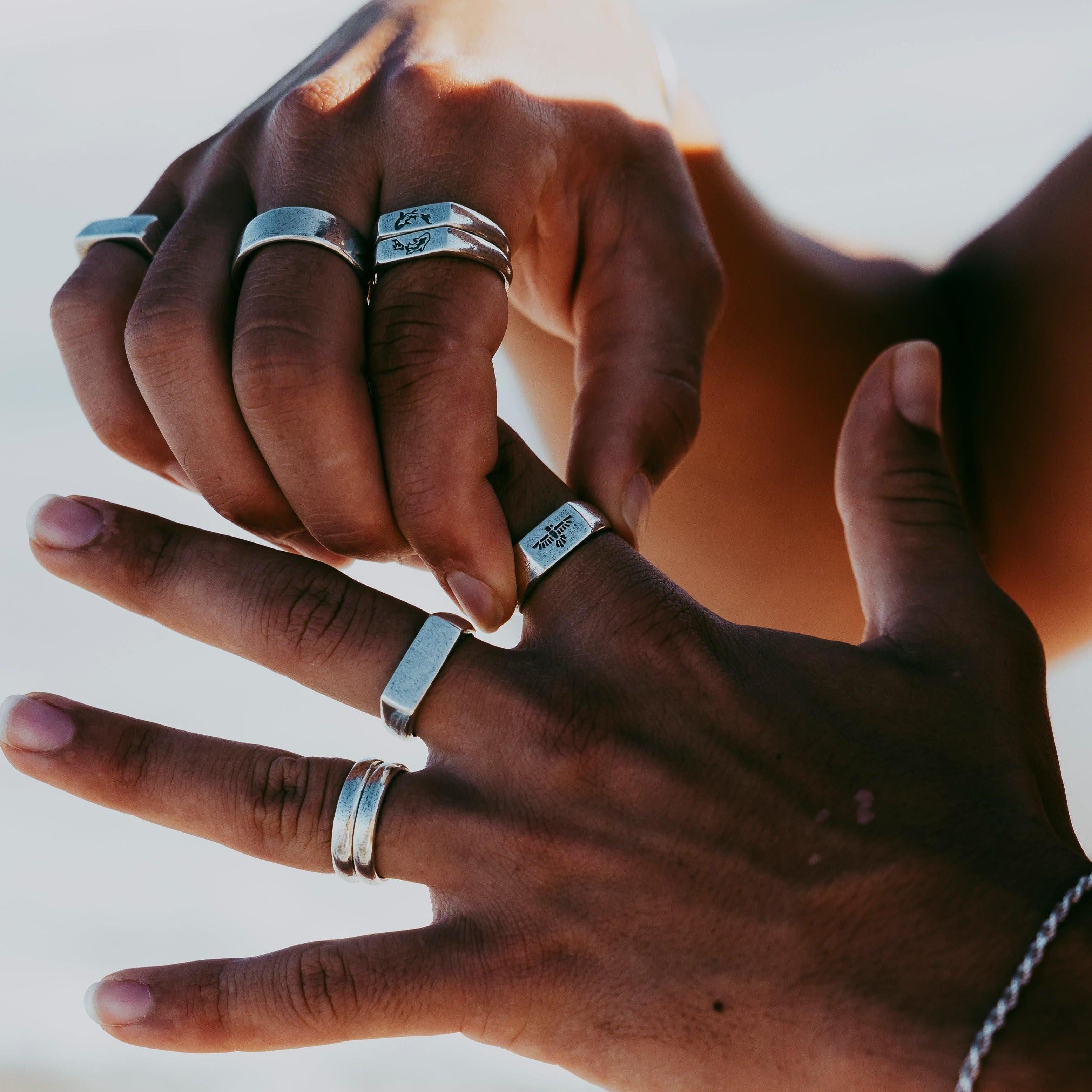 Two hands with medium brown skin tone wearing multiple silver rings and a silver bracelet, posed against a blurred light background, possibly outdoors in natural light. v2