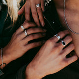 Close-up of two people wearing silver rings and necklaces, gently touching each others hands and chest, dressed in dark shirts with sunlight highlighting their skin and jewelry.