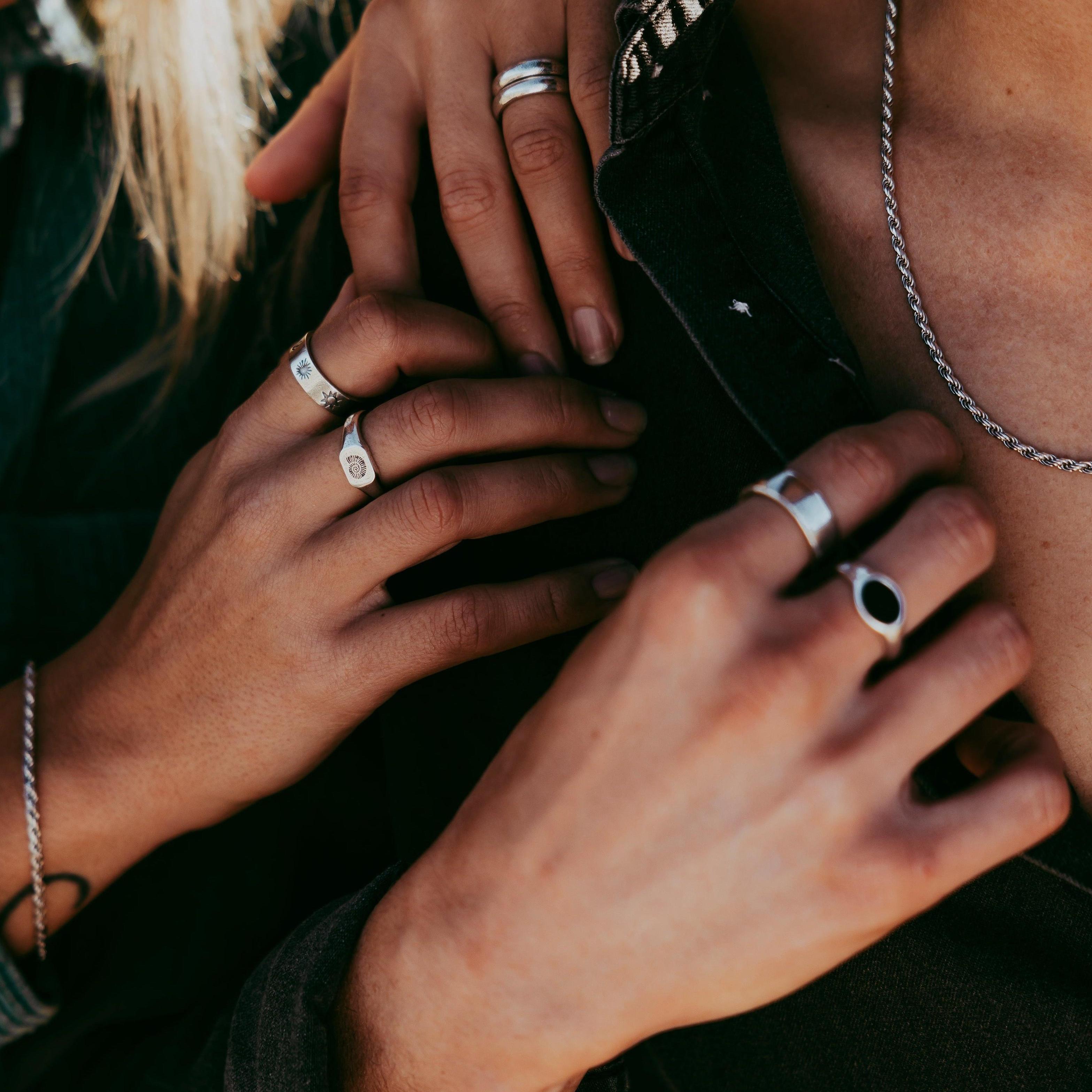 Close-up of two people wearing silver rings and necklaces, gently touching each others hands and chest, dressed in dark shirts with sunlight highlighting their skin and jewelry.