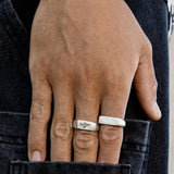 A close-up of a hand wearing two silver rings and a silver bracelet, with fingers resting on the back pocket of dark denim jeans. The hand is partially tucked into the pocket. v1