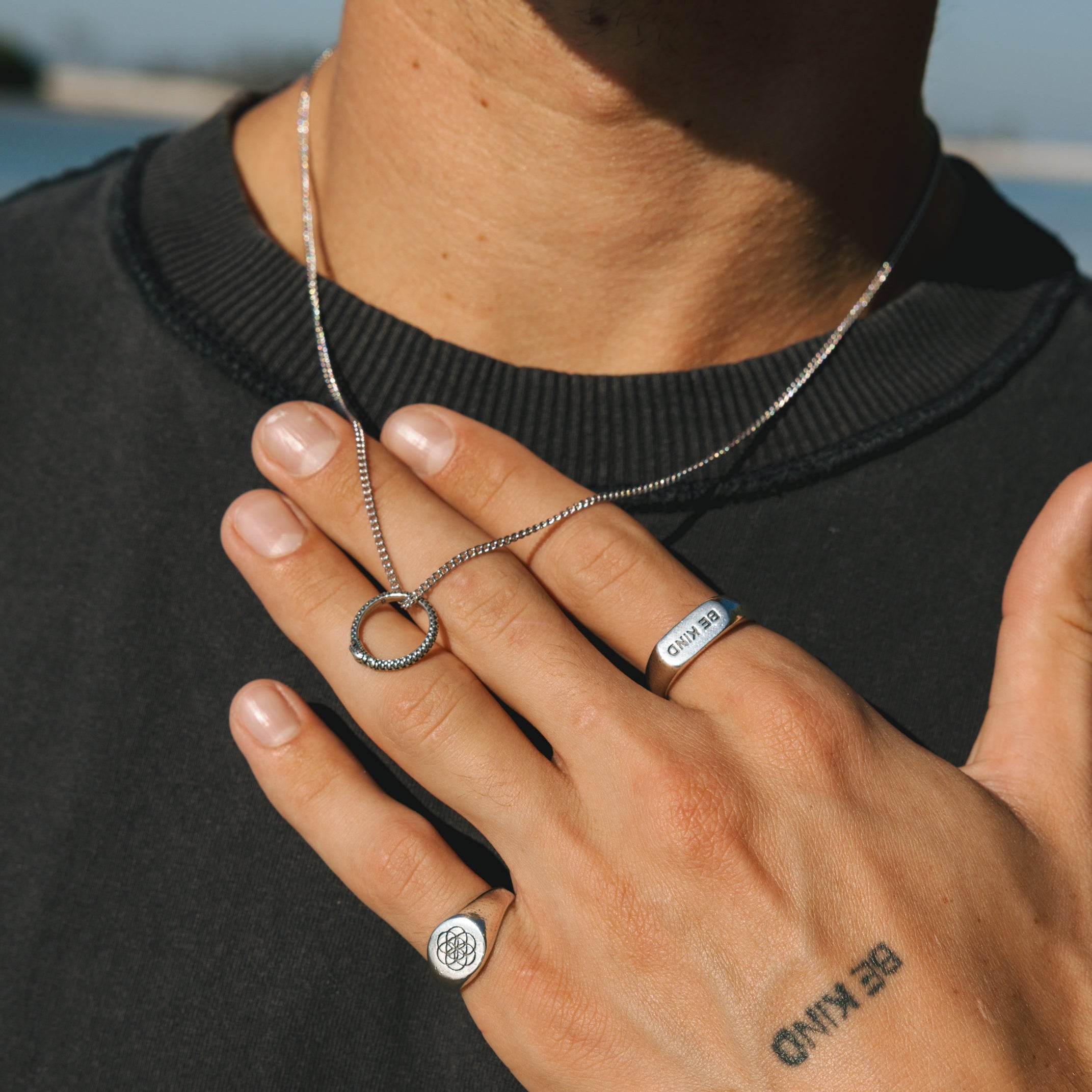 Person wearing a necklace and two rings with a tattoo on their hand, outdoors.