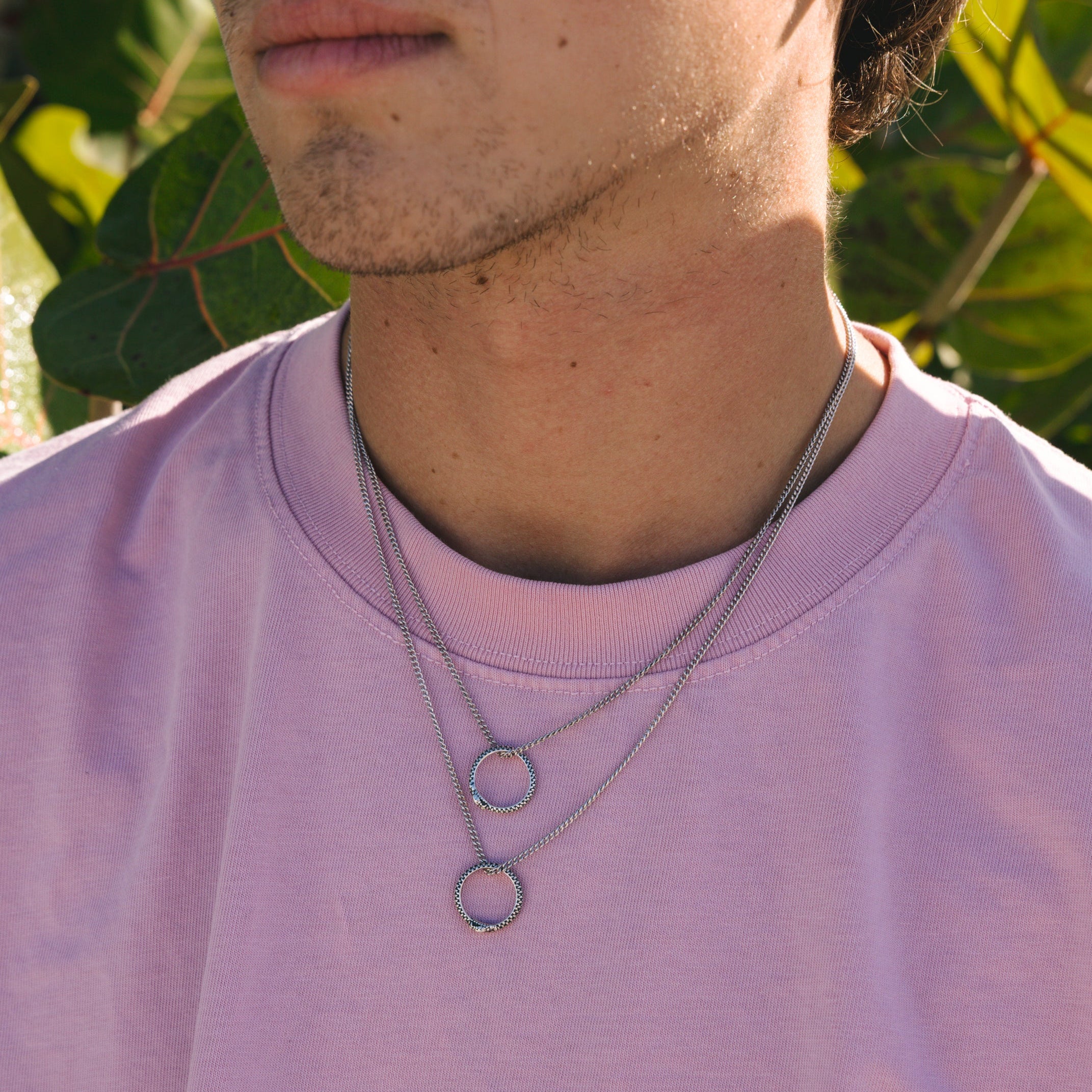 Person wearing a pink shirt with a necklace featuring a snake circular pendant, surrounded by green foliage.