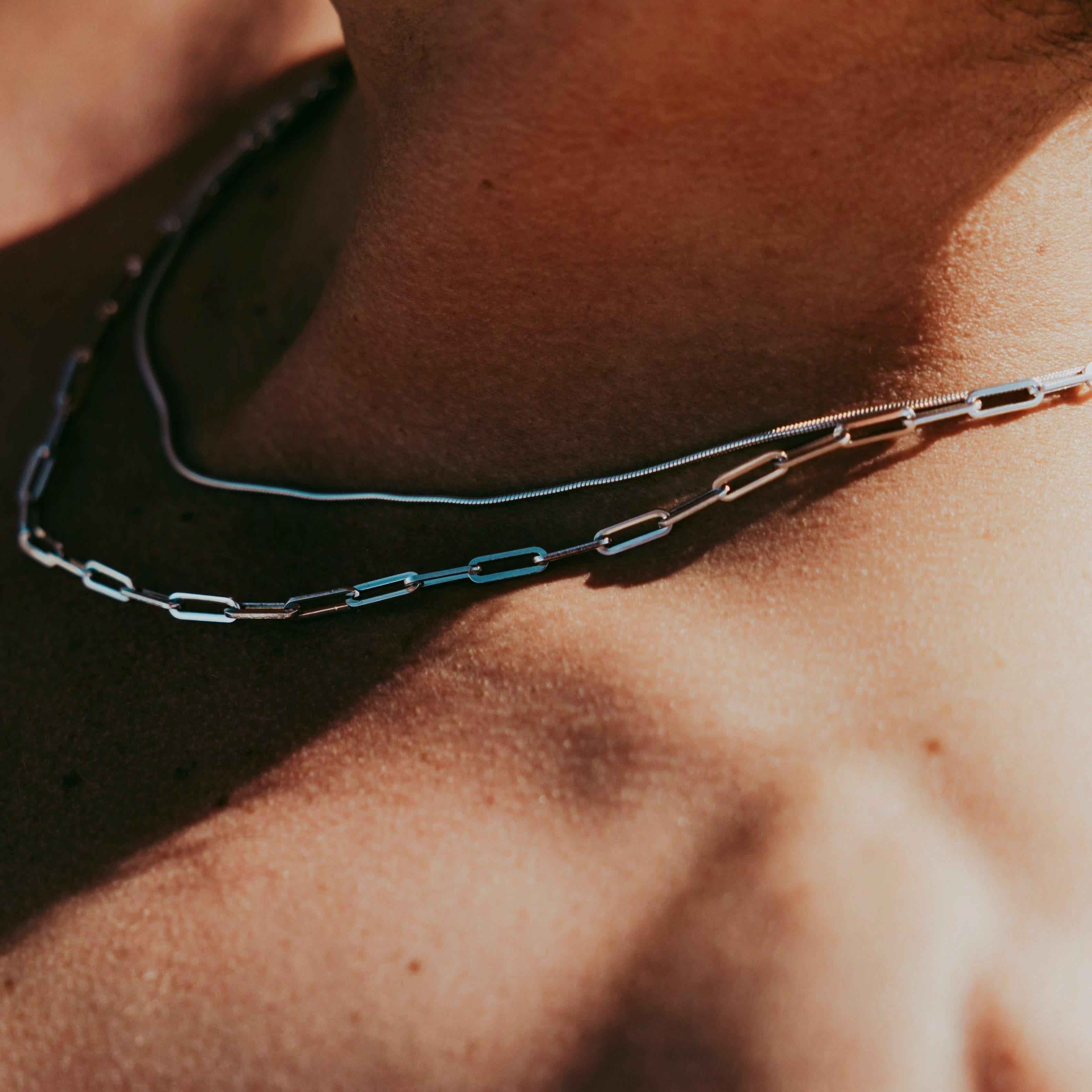 Close-up of a persons bare upper chest and neck wearing two silver chains, one with oval links and the other a smooth snake chain, lit by warm sunlight.